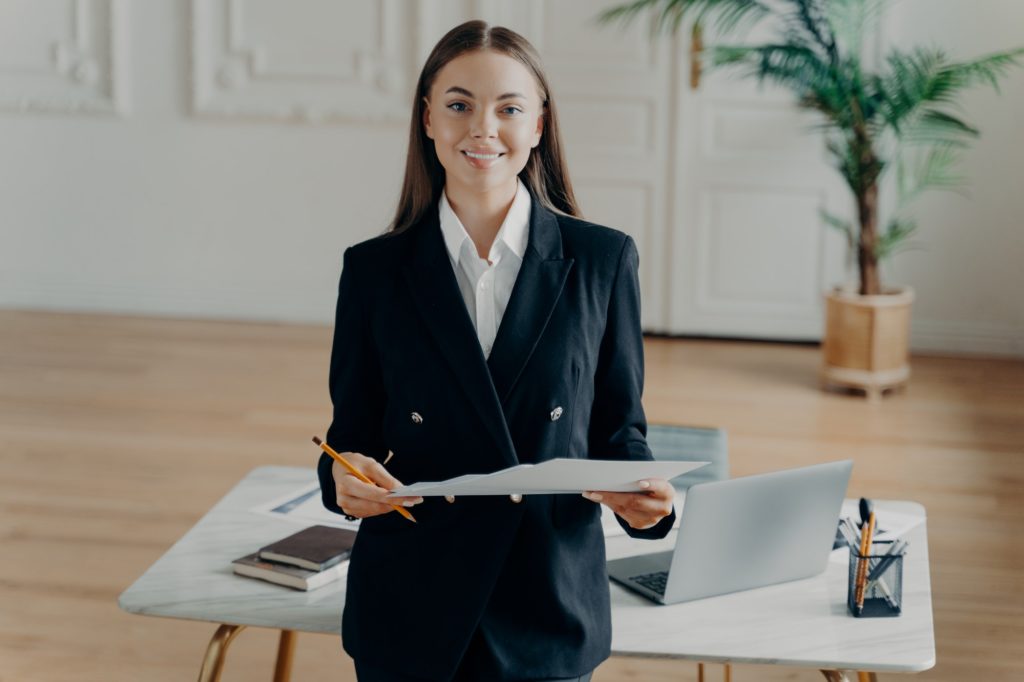 Young smiling bussiness woman looking at camera and holding document in front of office desk
